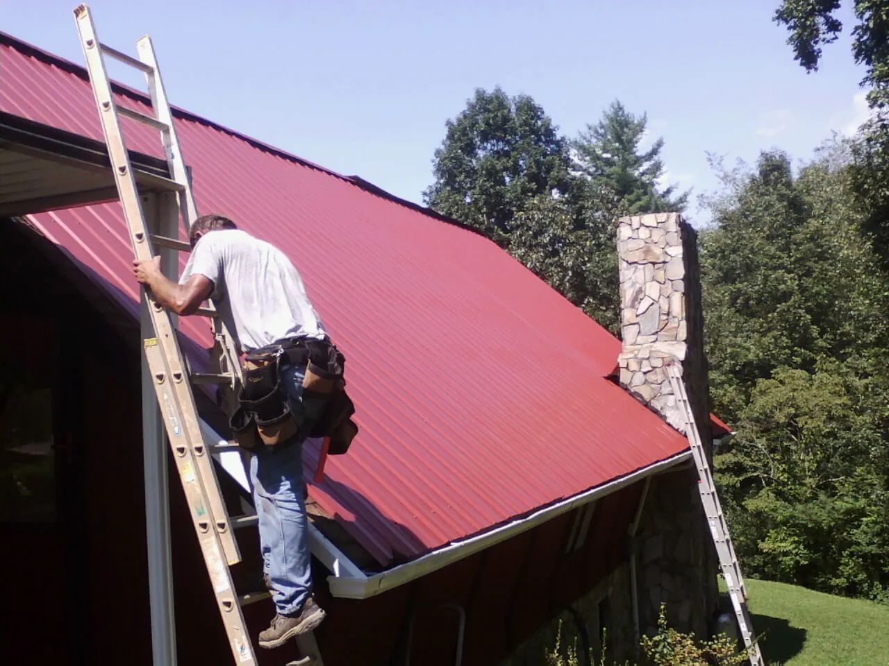 Roofer on a ladder beside a red metal roof and stone chimney — Soffit & Fascia Repair by Roofing Brothers in Oakman